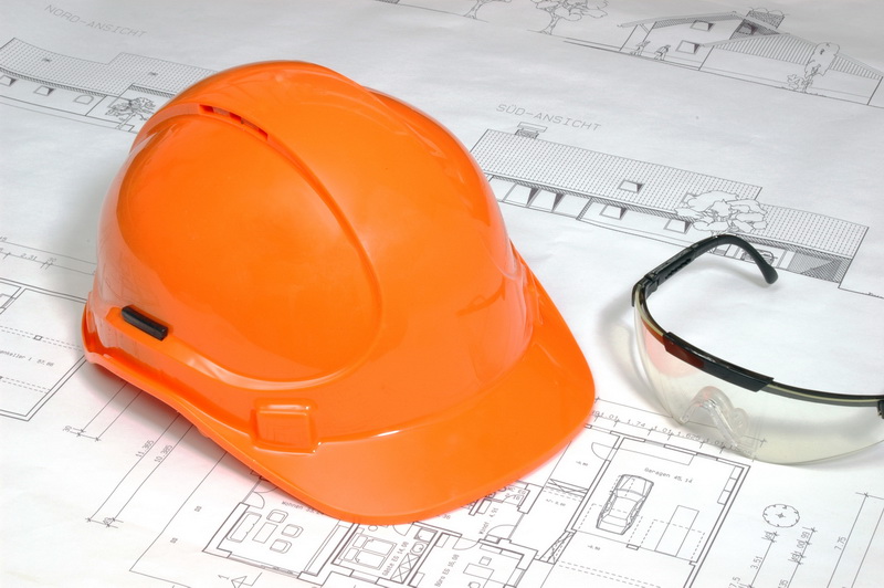 Ein orangener Bauhelm liegt auf einem Bauplan und daneben liegt eine Schutzbrille. 
